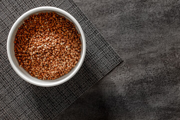 Raw buckwheat in bowl, on dark tablecloth background, top view, space to copy text