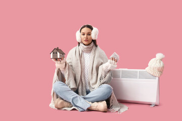 Young woman with house model, money and radiator on pink background. Price rise concept
