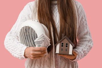 Young woman with electric fan heater, money and house model on pink background, closeup. Price rise...