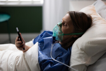 Elderly patient in hospital ward, oxygen mask on, using the phone to stay connected with family.