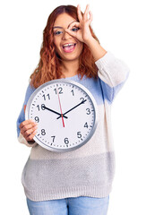 Young latin woman holding big clock smiling happy doing ok sign with hand on eye looking through fingers