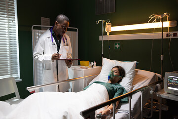 African doctor stands beside the unconscious girl in the hospital room, holding a folder and taking...