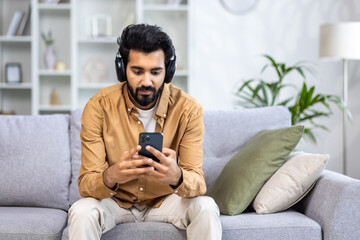 Obraz premium A young Indian man in headphones holds a phone in his hands, sits on a sofa in a bright living room, listens to music, enjoys rest.