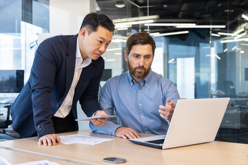 Two businessmen in suits work in the office, look at a laptop, discuss work issues, have a...