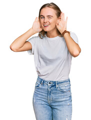 Beautiful young blonde woman wearing casual white t shirt trying to hear both hands on ear gesture, curious for gossip. hearing problem, deaf