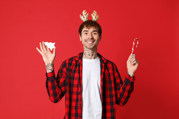 Tattooed young man in reindeer horns with Christmas tree toy and candy cane on red background