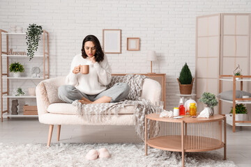 Sick young woman with cup of hot tea sitting on sofa at home
