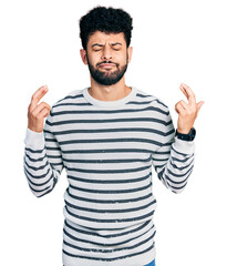 Young arab man with beard wearing casual striped sweater gesturing finger crossed smiling with hope and eyes closed. luck and superstitious concept.