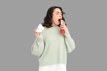 Sick young woman with spray for sore throat and tissue on grey background