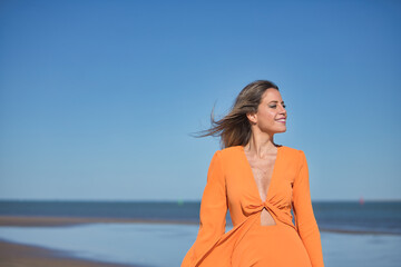 Young and beautiful woman in an orange dress walking on the beach, happy and relaxed, with the wind moving her dress and hair. Concept beauty, fashion, trend.