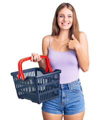 Young beautiful blonde woman holding supermarket shopping basket pointing finger to one self smiling happy and proud