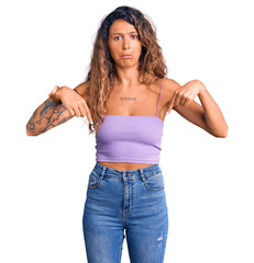 Young hispanic woman with tattoo wearing casual clothes pointing down looking sad and upset,...