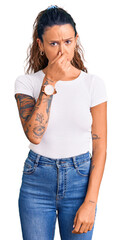 Young hispanic woman with tattoo wearing casual white tshirt smelling something stinky and disgusting, intolerable smell, holding breath with fingers on nose. bad smell