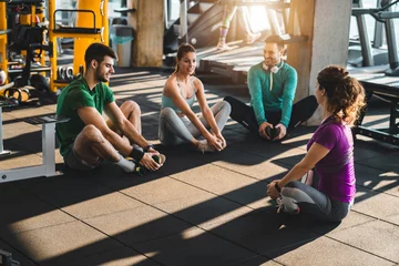 Poster Im Rahmen Group of diverse athletic people exercising in a health club. Young friends doing stretching exercises while sitting on the floor in gym. Fitness instructor exercising with a group of three people. © Dorde