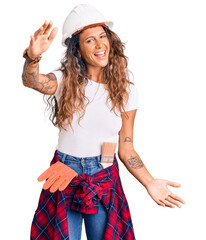 Young hispanic woman with tattoo wearing hardhat and builder clothes looking at the camera smiling with open arms for hug. cheerful expression embracing happiness.