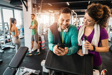 Happy athletic couple using phone while taking a break from weight training in the gym. Young people talking between exercises in fitness center.