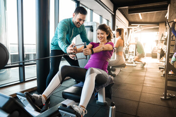 Happy athlete doing rowing workout with personal trainer in the gym. Dedicated fitness instructor...