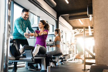 Happy athlete doing rowing workout with personal trainer in the gym. Dedicated man coach showing right exercise technique to young female newcomer.