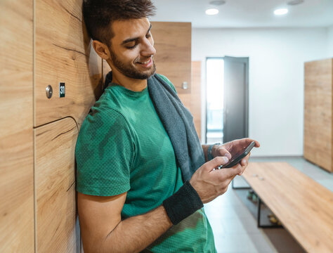 Happy athletic man standing in locker room while using mobile phone and taking a break from exercising. Smiling guy checking notifications on his cellphone.