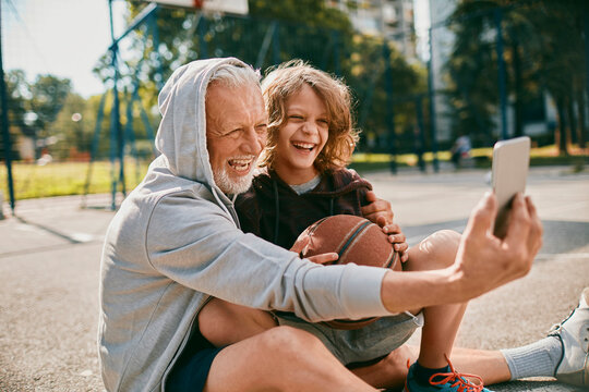 Grandfather and grandson taking a selfie on a smart phone while resting from playing basketball outdoors on a basketball court in the city