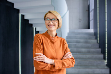 Portrait of a smiling businesswoman in glasses, standing with her arms folded on her chest, smiling and looking away.