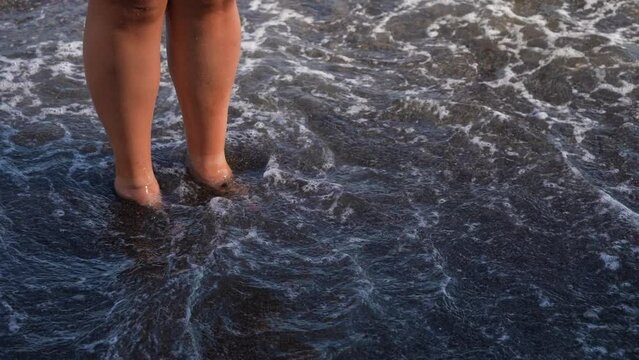 Close-up cropped shot legs of unrecognizable woman walking along wave of sea water and sand on summer beach. Concept of relax, travel, freedom and vacation. Shooting in slow motion.