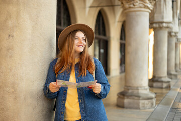 Fototapeta na wymiar Pretty young woman staying on the street holding paper map in hands. 30s girl in hat posing on Market Square in Krakow. Traveling Europe, High quality photo, The Cloth Hall, Vacation concept