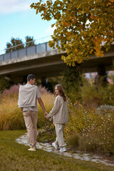 Fototapeta premium Young couple in love walking in the autumn park holding hands looking in the sunset. Loving couple holding hands while walking at sunset. The hands of the male and female lovers who hold hands walk