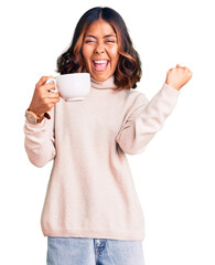Young beautiful mixed race woman holding a cup of coffee screaming proud, celebrating victory and...