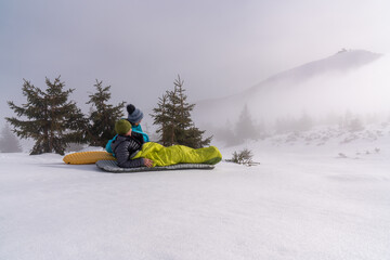 A middle-aged woman and a man are sitting in sleeping bags on mattresses in the snowy mountains....