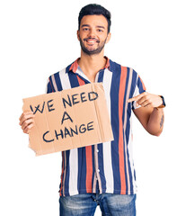 Young handsome hispanic man holding we need a change banner smiling happy pointing with hand and...