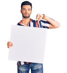 Young handsome hispanic man holding cardboard banner with blank space with angry face, negative...