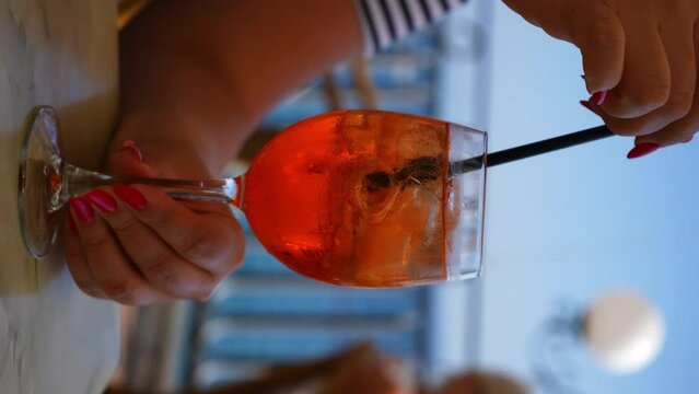 Vertical cropped shot of unrecognizable woman stirring aperol spritz cocktail with straw and drinking outdoors cafe on summer day on background of sea. Delicious alcoholic drink with citrus fruits.