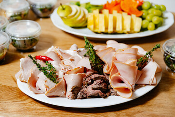 Fototapeta na wymiar Assorted Cold Meat Platter with Fresh Fruit. A variety of sliced cold meats served on a platter, accompanied by herbs and chili peppers, fresh fruit platter on a background.