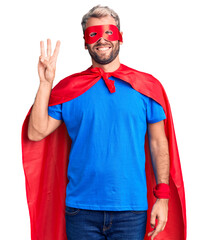 Young blond man wearing super hero custome showing and pointing up with fingers number three while smiling confident and happy.