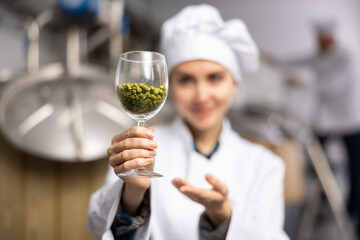 Woman brewmaster in whihte coat holding glass with hops pellets.