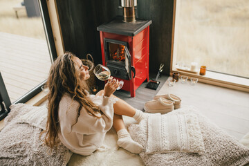 Young woman sitting by the fireplace in white sweater, drinking wine in cozy log cabin.