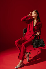 Fashionable confident woman wearing trendy red suit blazer, classic trousers, metallic silver color shoes, holding black tweed  bag, sitting on chair, posing on red background. Studio fashion portrait