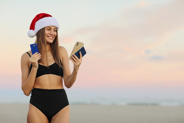 Happy slim young woman in Santa Claus hat, bikini outdoors shows smartphone, credit card, passport for book online ticket to travel on Christmas and New Year 2024 holiday vacation at tropical resort