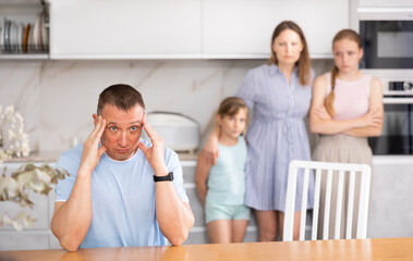 Fototapeta premium Upset and puzzled man sitting at kitchen table, deep in thought, experiencing some serious problem or stressful situation. Wife and two teenage daughters standing on blurred background