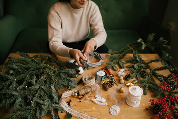 Woman making Christmas wreath using natural pine branches, sitting near wooden table.