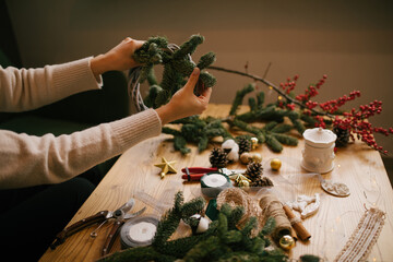 Woman making Christmas wreath using natural pine branches, sitting near wooden table.