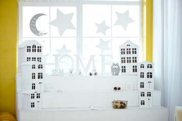 Beautiful decorative area near the window with white small houses, the inscription HOME, stars, and the moon. Steps for placing home New Year's decor. Cozy home space. Interior design.