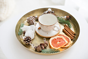 Christmas composition on a metal tray with a white cup on a saucer, pine cones, fir branches, cinnamon sticks, and dried oranges. DIY festive winter decoration ideas. New Year celebration decor.