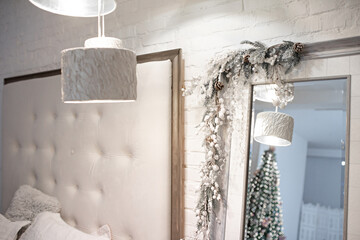 A mirror in a bright modern bedroom decorated with a snow-covered fir branch garland reflects a Christmas tree with twinkling lights. A soft headboard of the bed and a contemporary lamp.