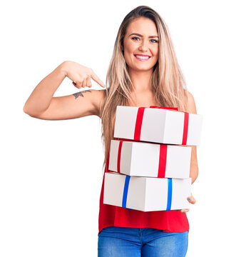 Young beautiful blonde woman holding birthday gifts pointing finger to one self smiling happy and proud