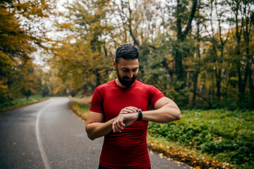 Fototapeta premium A runner is standing on road in nature and looking at smartwatch.