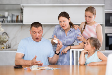 Obraz na płótnie Canvas Upset puzzled man counting banknotes at kitchen table while disgruntled wife and two teenage daughters demanding for pocket money