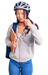 Young beautiful chinese girl wearing student backpack and bike helmet holding book annoyed and frustrated shouting with anger, yelling crazy with anger and hand raised