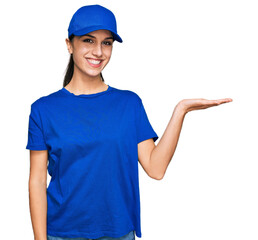 Young hispanic girl wearing delivery courier uniform smiling cheerful presenting and pointing with palm of hand looking at the camera.